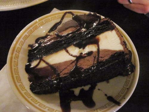 A slice of layered chocolate cake with cream and chocolate sauce drizzled on top, served on a decorative plate.