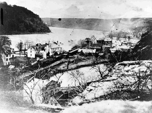 Historic black-and-white photo of a riverside town with houses and trees, surrounded by hills and water.