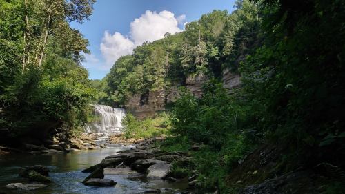 A serene waterfall cascades into a rocky stream, surrounded by lush greenery and steep cliffs under a blue sky.