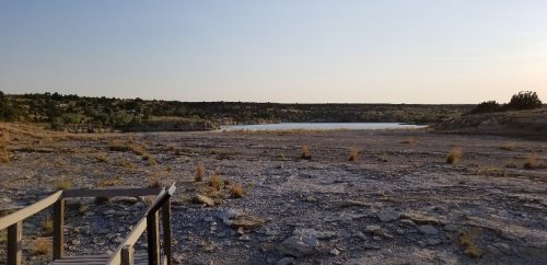A dry, rocky landscape with sparse vegetation and a distant lake under a clear sky.