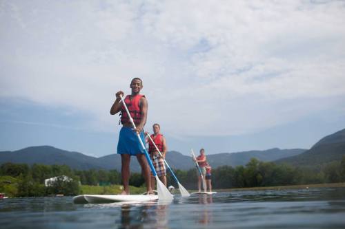 Three people paddleboarding on a calm lake, surrounded by mountains and greenery, under a partly cloudy sky.