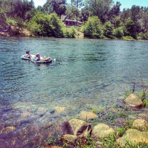 Two people relax on inflatable rafts in a calm river, surrounded by lush greenery and rocky riverbank.