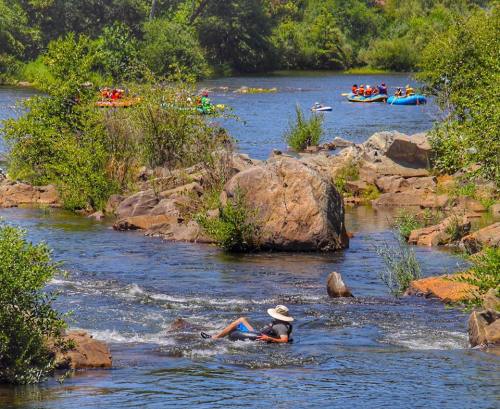 A person relaxes in a river while kayakers paddle in the background, surrounded by lush greenery and rocky terrain.