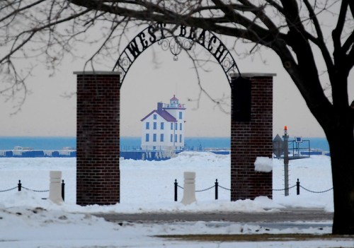 A snowy landscape framed by an archway, featuring a distant white lighthouse by the lake under a gray sky.