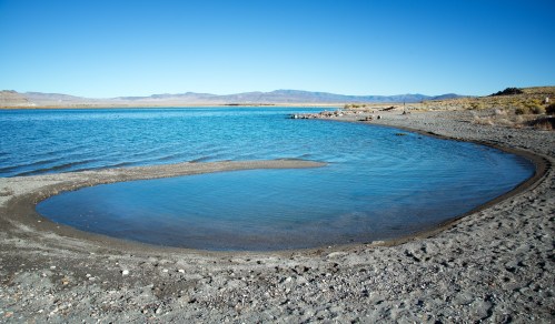A serene lake with clear blue water, sandy shore, and distant mountains under a bright blue sky.