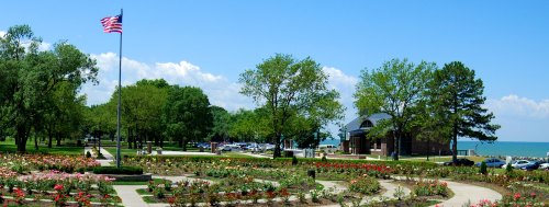 A scenic park with a flagpole, rose garden, trees, and a view of the lake under a clear blue sky.