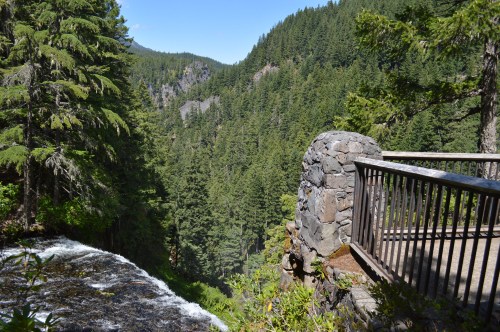 A scenic view of a lush green valley with a waterfall, seen from a stone overlook surrounded by trees.