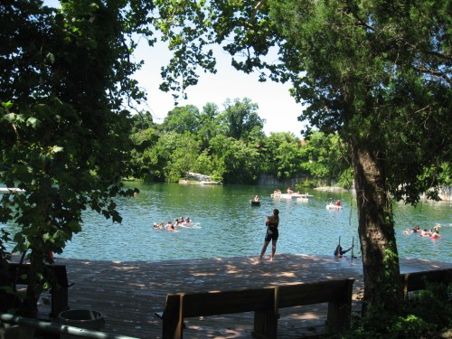 A serene lake scene with people swimming and relaxing on a wooden dock surrounded by lush greenery.