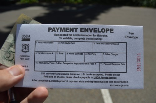 A hand holds a payment envelope with instructions and a dollar bill, set against a blurred outdoor background.