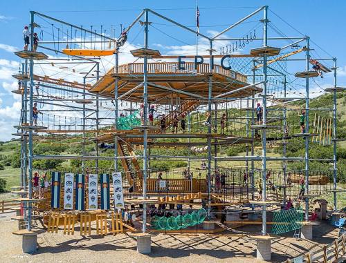 A large outdoor climbing structure with multiple levels, ropes, and platforms, surrounded by greenery and blue skies.