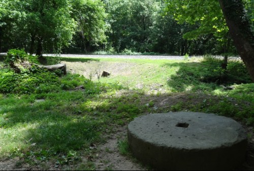 A grassy area with a large stone wheel and trees, near a road in a wooded setting.