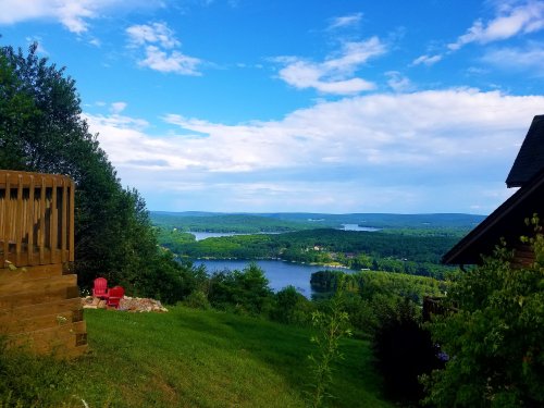 Scenic view of a lake surrounded by greenery under a blue sky with clouds, featuring red chairs on a hillside.