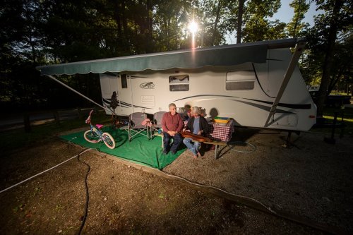 A couple sits outside their camper in a wooded area, with a bicycle and chairs nearby, enjoying a sunny day.