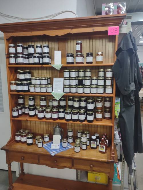 A wooden shelf displaying various jars of preserves and sauces, organized neatly with labels and a small table underneath.