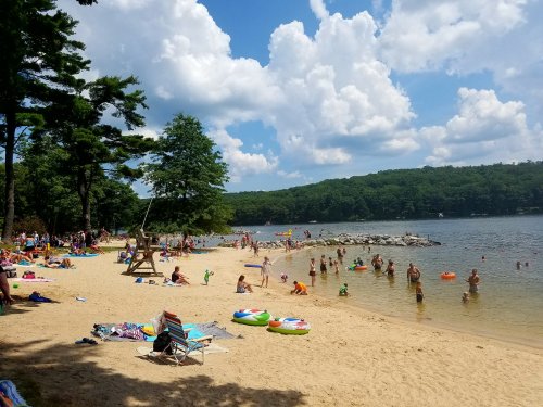 A busy beach scene with people swimming, lounging on the sand, and enjoying the water under a partly cloudy sky.