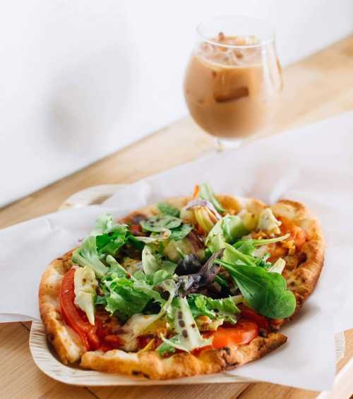 A wooden plate with a fresh salad-topped pizza and a glass of iced coffee on a light wooden table.