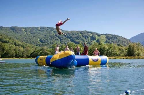 A person jumps off a water trampoline while others watch from the inflatable structure in a scenic lake setting.
