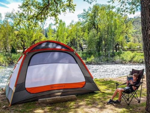 A boy sits in a chair by a river, reading, next to a colorful tent surrounded by trees and greenery.