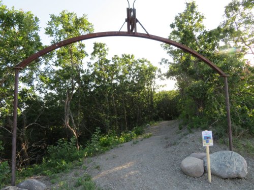 A metal archway marks the entrance to a wooded path, surrounded by greenery and a sign on a post nearby.