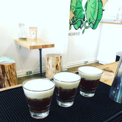 Three coffee drinks with frothy tops sit on a bar counter, with wooden stools and a green mural in the background.