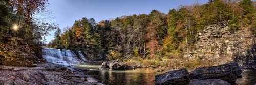 A panoramic view of a waterfall surrounded by autumn foliage and rocky terrain under a clear blue sky.