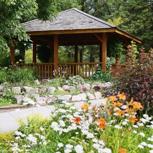 A wooden gazebo surrounded by vibrant flowers and greenery in a peaceful garden setting.