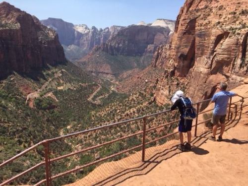 Two hikers stand by a railing, overlooking a vast canyon with red rock formations and green vegetation below.