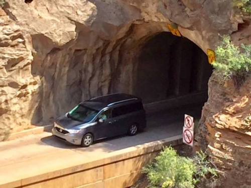 A black minivan drives through a rocky tunnel with warning signs nearby.