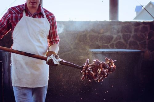 A person in an apron holds a skewer of grilled meat over a smoky outdoor grill.
