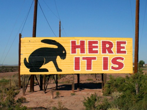 A large yellow billboard features a black rabbit silhouette and the text "HERE IT IS." Set against a clear blue sky.