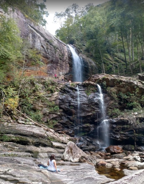 A serene waterfall cascades down rocky cliffs, surrounded by lush greenery, with a person sitting on the rocks below.