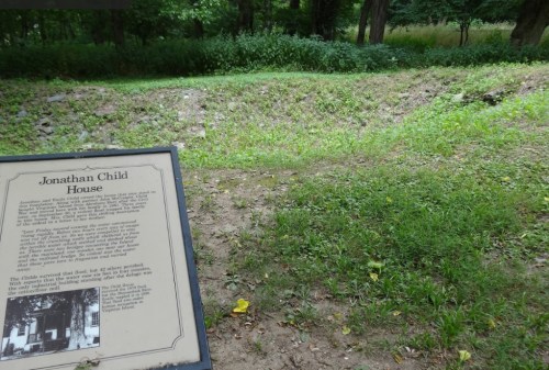 A grassy area with a sign about the Jonathan Child House, surrounded by trees and overgrown vegetation.