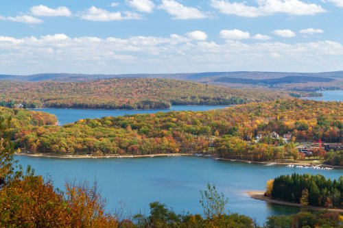 A scenic view of a lake surrounded by colorful autumn foliage and rolling hills under a partly cloudy sky.