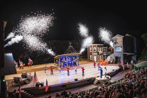 A lively outdoor performance with dancers, fireworks, and an audience, set in a western-themed venue at night.