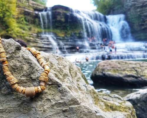 A close-up of a beaded necklace on a rock, with a waterfall and people swimming in the background.