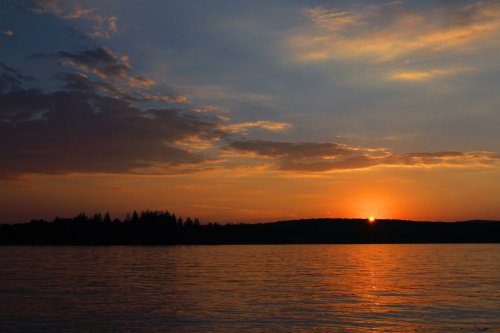 Sunset over a calm lake, with vibrant orange and blue hues reflecting on the water and silhouetted trees in the background.