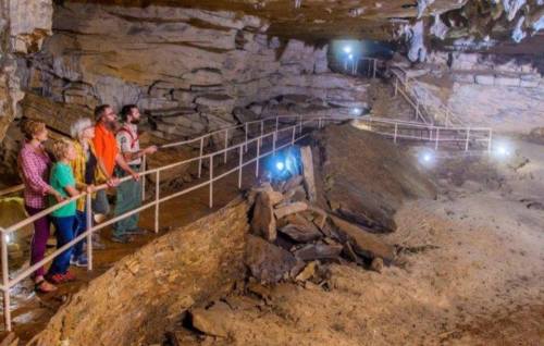 A group of visitors stands on a walkway in a cave, observing the rock formations and illuminated areas around them.