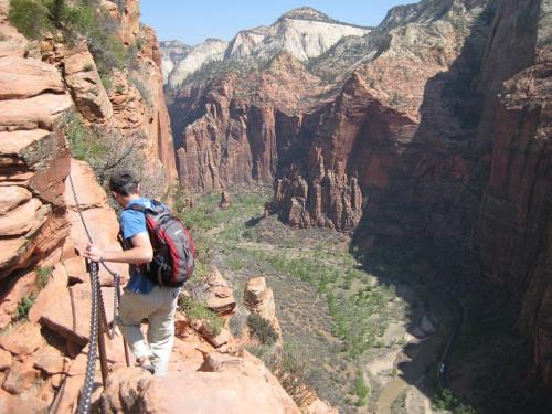 A hiker navigates a rocky trail overlooking a deep canyon with lush greenery and a river below.