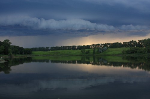A serene lake reflects dark storm clouds and lush green hills under a moody sky at dusk.
