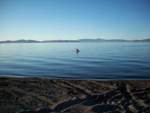 A calm lake with a person kayaking in the distance, surrounded by mountains under a clear blue sky.