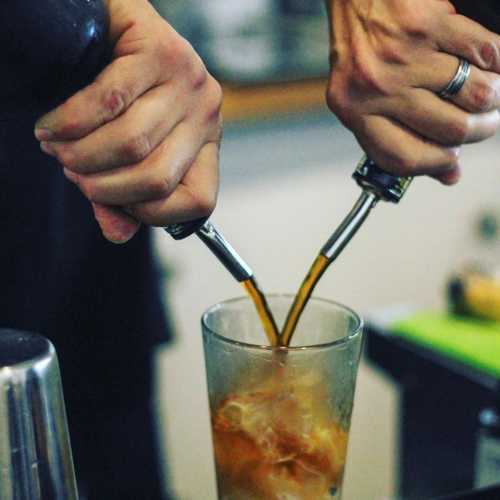 A close-up of hands pouring a dark liquid into a glass, with a blurred background of a bar or kitchen setting.