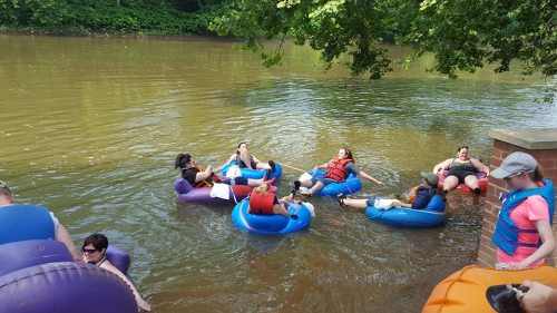 People relaxing on colorful inner tubes in a river, surrounded by greenery on a sunny day.