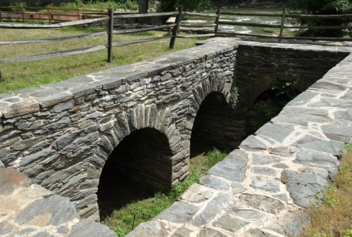 A stone bridge with three arches spans a grassy area near a river, surrounded by a wooden fence.