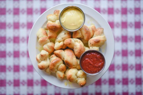 A plate of golden-brown breadsticks served with two dipping sauces: yellow and red, on a checkered tablecloth.