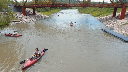 Three kayakers paddle on a calm river beneath a red metal bridge, surrounded by greenery and rocky banks.