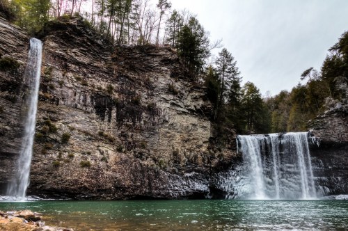 Two waterfalls cascade down rocky cliffs into a serene green pool, surrounded by trees and rugged terrain.