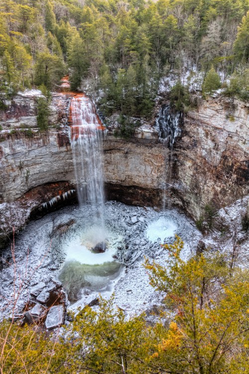 A stunning waterfall cascades into a rocky pool, surrounded by snow and lush green trees in a serene natural setting.