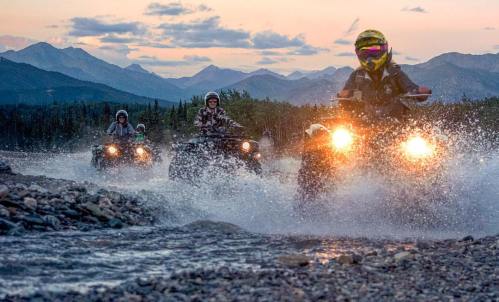 Four riders on ATVs splash through a stream, with mountains and a sunset in the background.