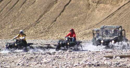 Three off-road vehicles splash through shallow water on a rocky terrain, surrounded by dry, mountainous landscape.
