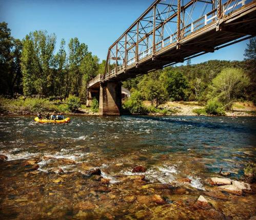 A group rafting on a river beneath a metal bridge, surrounded by lush greenery and rocky riverbed.